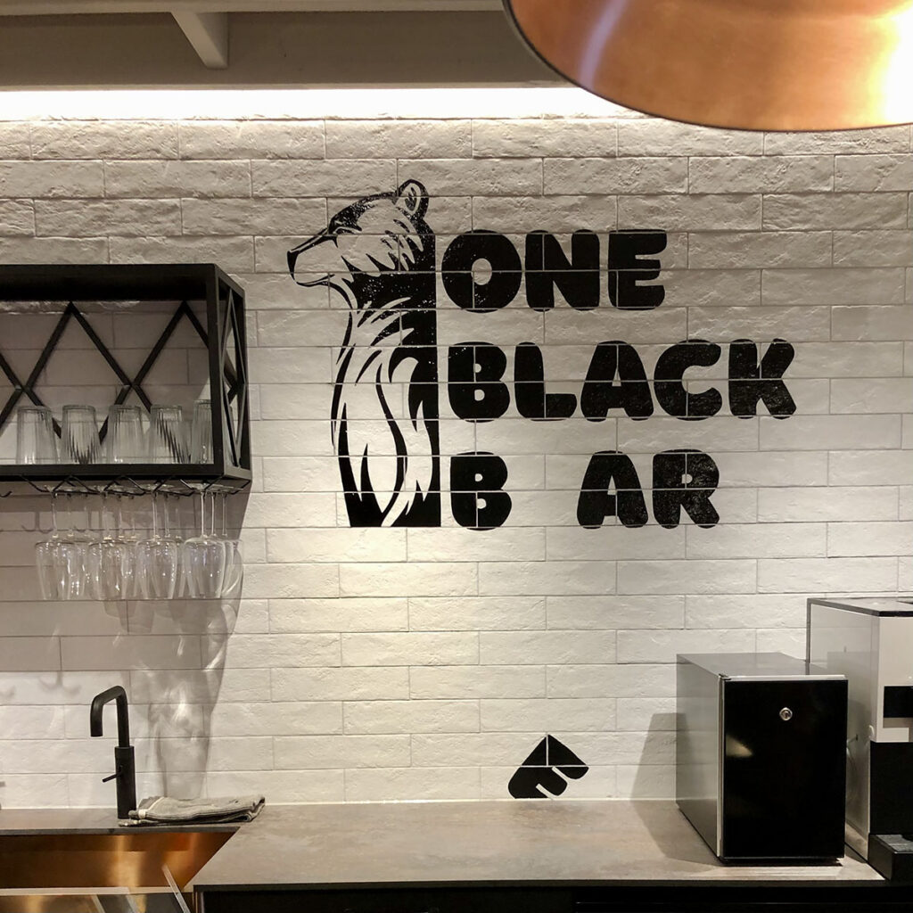 Hand-painted black signwriting and bear mural on white tiled wall inside One Black Bear in Birmingham, featuring bold lettering and clean brushwork for a modern interior finish.