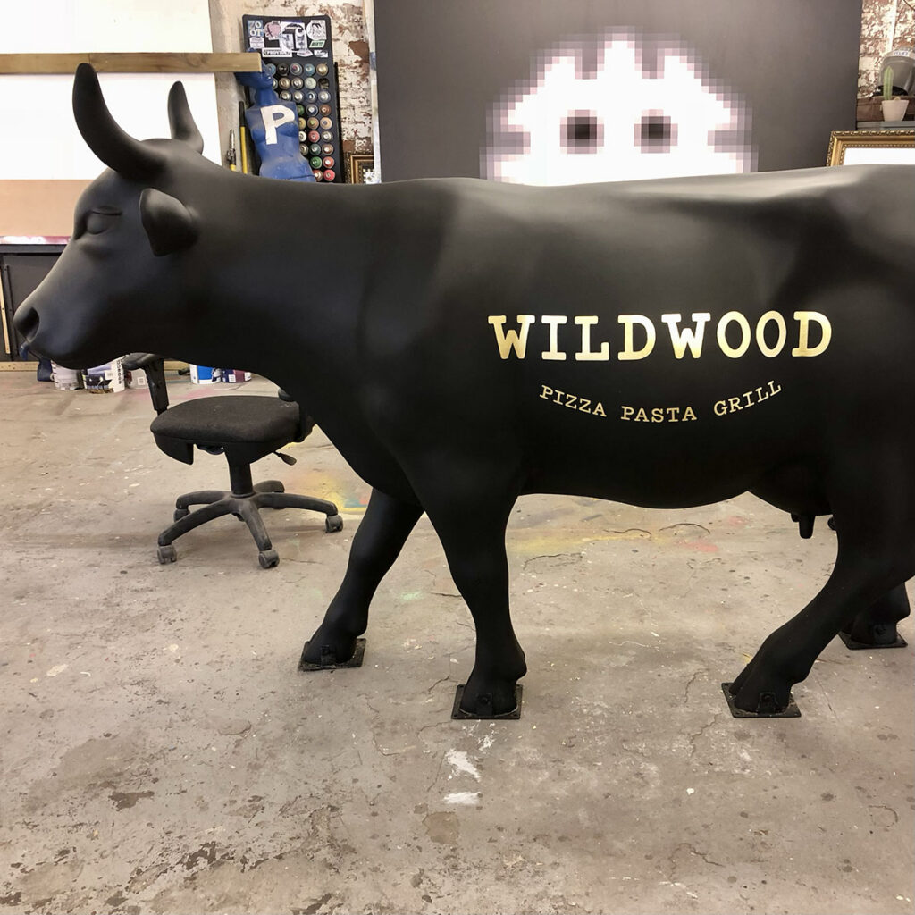 Hand-painted gold lettering reading “Wildwood Pizza Pasta Grill” on a matte-black life-size cow sculpture, completed in a workshop setting.