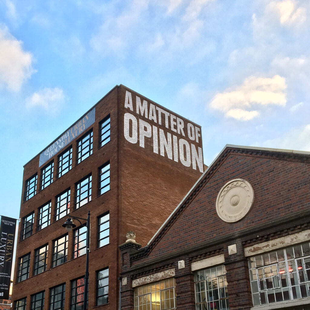 Large hand-painted mural reading “A MATTER OF OPINION” across the top of a Jewellery Quarter building in Birmingham, adding bold character to the historic red-brick skyline.
