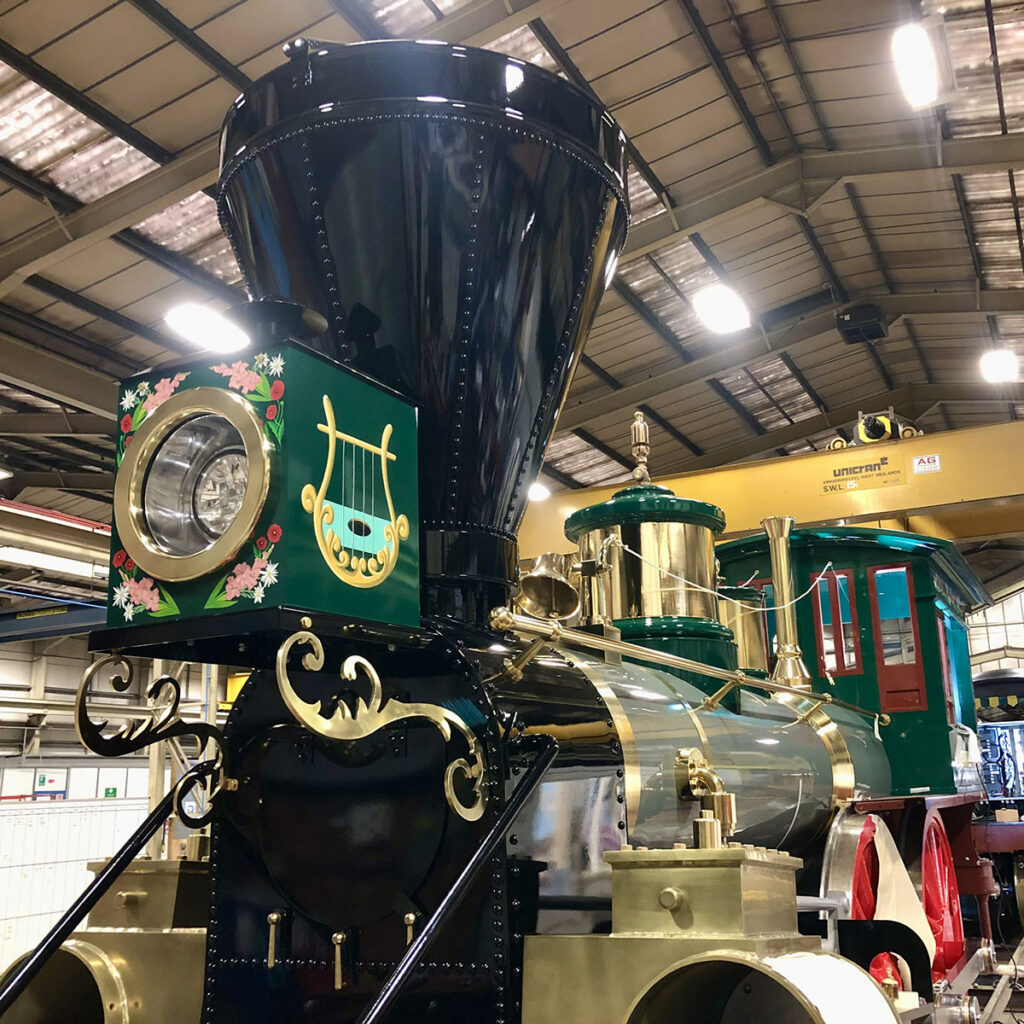 Hand-painted detailing on an electric train built in the style of a classic steam locomotive, featuring floral artwork, scrolls and a gold lyre emblem across the front panels.