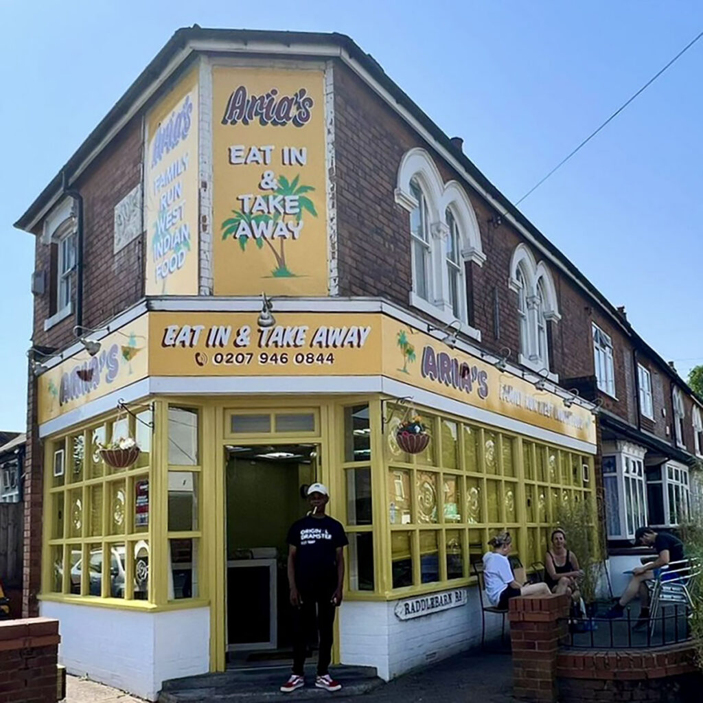 Hand-painted shop frontage for Aria’s, a fictional West Indian takeaway created for BBC One and Netflix’s Champion, featuring bold yellow signage and characterful lettering on a corner building in Birmingham.