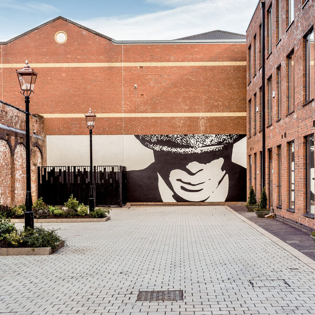 Hand-painted Peaky Blinders–inspired mural in a Birmingham courtyard, featuring a bold black-and-white portrait tucked beneath the brickwork of a new development.
