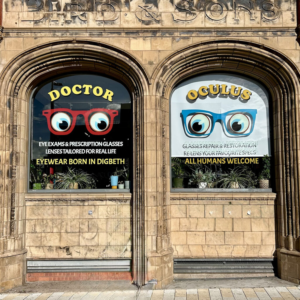 Hand-painted window lettering for Doctor Oculus in Digbeth, featuring bold illustrated glasses and clean signwritten text across two large arched shopfront windows of the Zellig Building