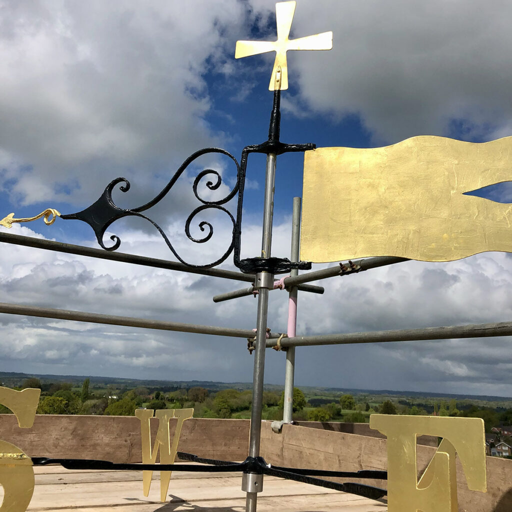 23.5-carat gold leaf applied to a restored weather vane, catching the light high above the rooftops while lightning technicians and steeplejacks carry out the installation work.