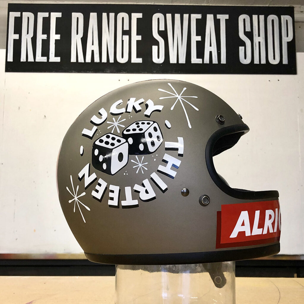 Hand-painted motorcycle helmet featuring retro dice artwork, “Lucky Thirteen” lettering and mid-century starbursts, photographed in the studio against a bold typographic backdro