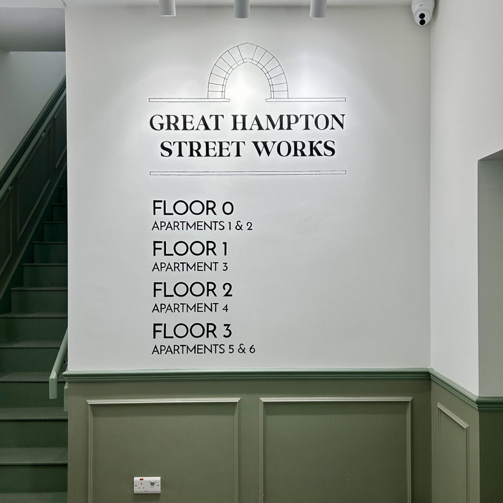 Hand painted apartment directory at Great Hampton Street Works in Birmingham, wayfinding signwriting listing floors and apartments on a clean white lobby wall beside a green staircase.