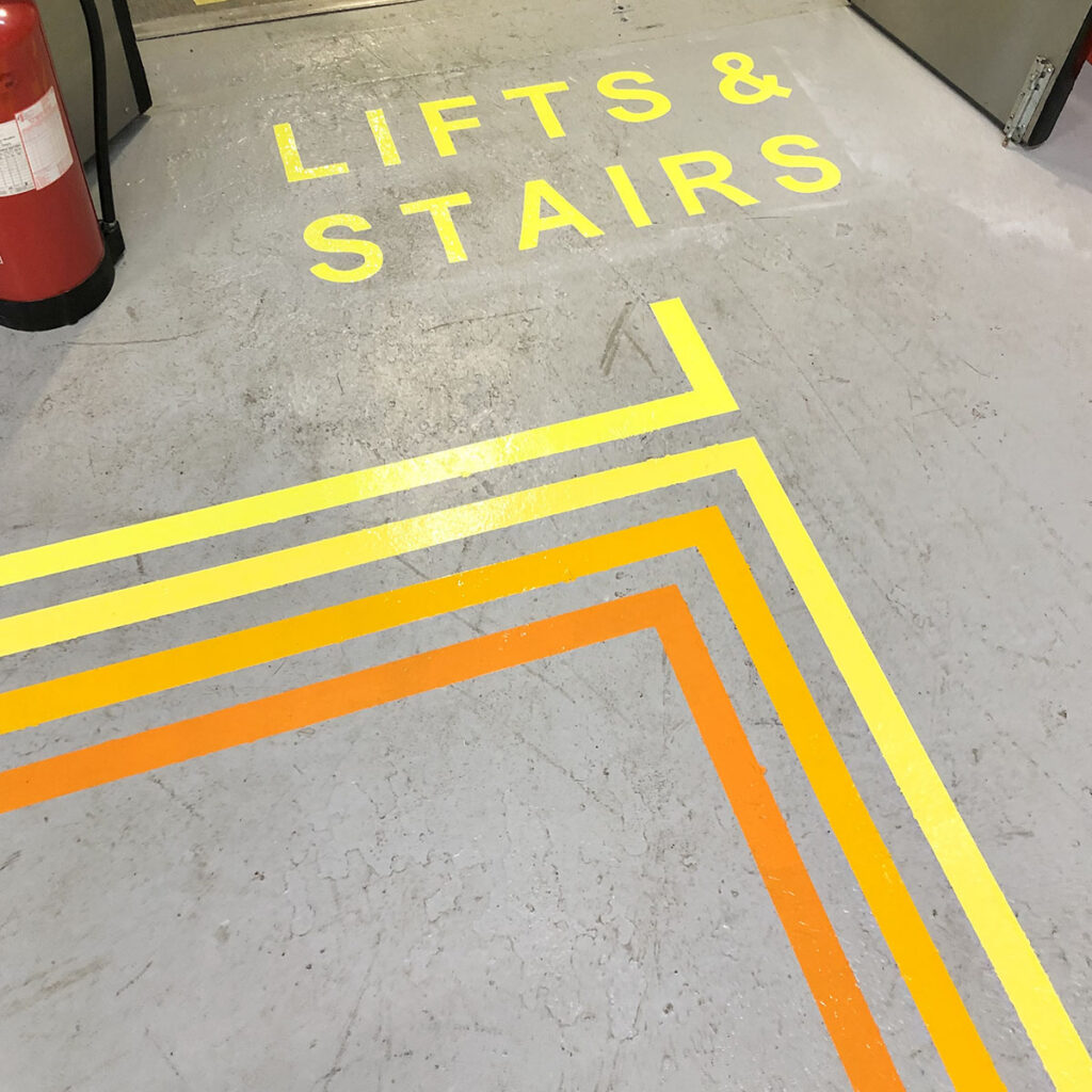 Hand-painted wayfinding on a concrete floor, with yellow and orange directional lines leading visitors toward lifts and stairs in a busy workspace.