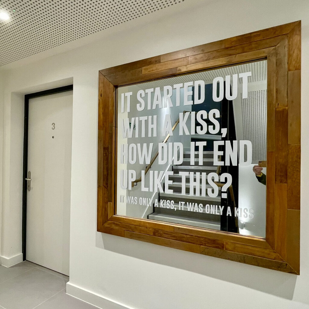 Hand-painted white lettering on a large framed mirror inside a modern apartment hallway, featuring lyrics from Mr. Brightside by The Killers.