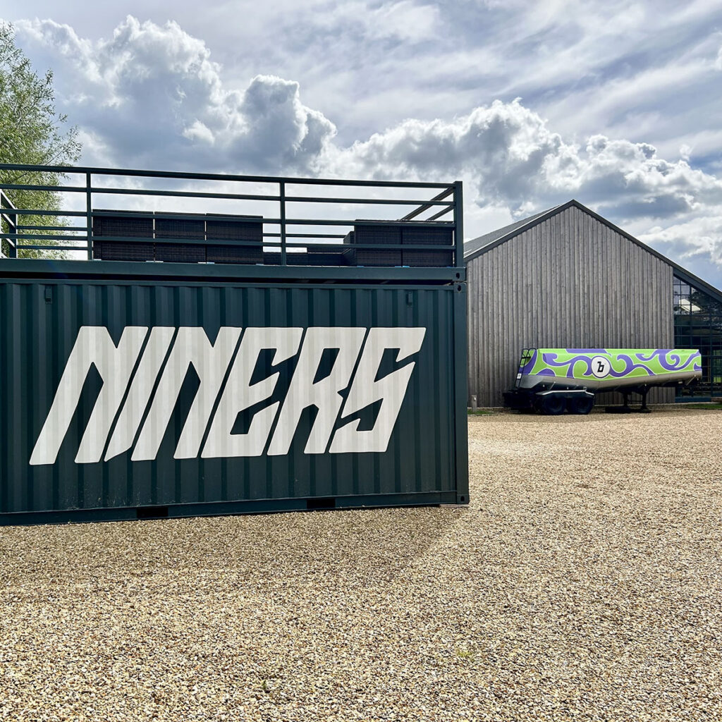 Hand-painted “NINERS” lettering on a dark green shipping container at Boxen­gasse, with bold angled type standing out against the rural yard and modern barn buildings in rural Oxfordshire.
