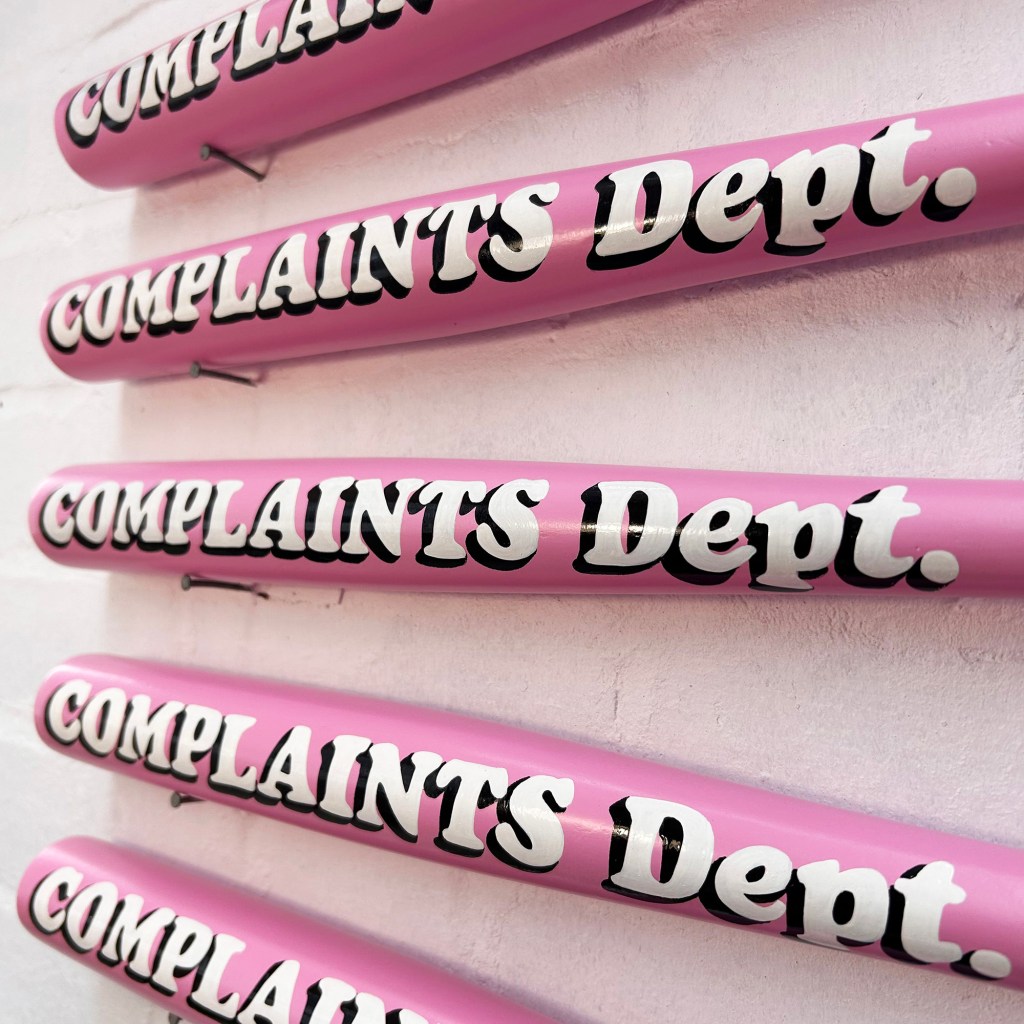 Close up of white hand lettered typography on pink Complaints Dept bat